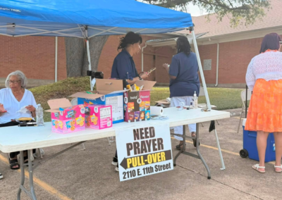 Members of the evangelism and outreach team sitting outside under a tent. A sign says drive-thru prayer.